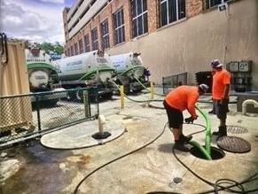 Two workers are pumping out a grease trap.  隔油池是人行道上的一个洞，有两根管子从那里出来.  这两根管子将烟雾从捕集器中吸出，并将其吸入停在捕集器后面的三辆卡车中.